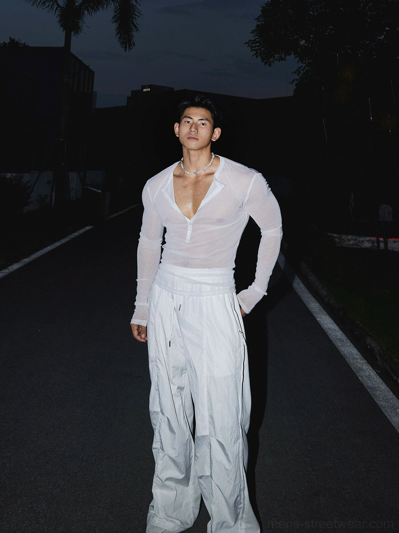 Full-length front view of a male model wearing a white sheer sparkly Henley top with wide-leg pants and partially open neckline