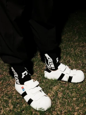 White shell-toe hidden-lift sneakers with black side stripes and velcro straps worn with black pants outdoors on grass