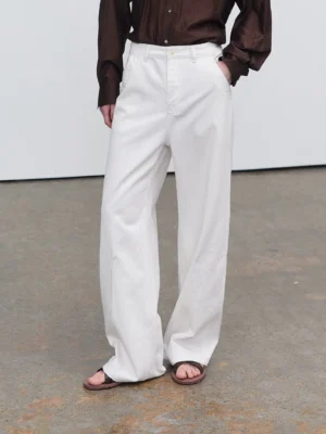 White slub textured relaxed straight leg jeans shown from a low front angle with a long stacked hem and slide sandals