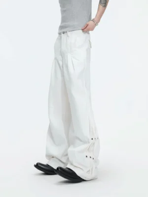 White snap detail draped wide leg pants shown in a low-angle side view with pleated high waist and adjustable studded hem tabs