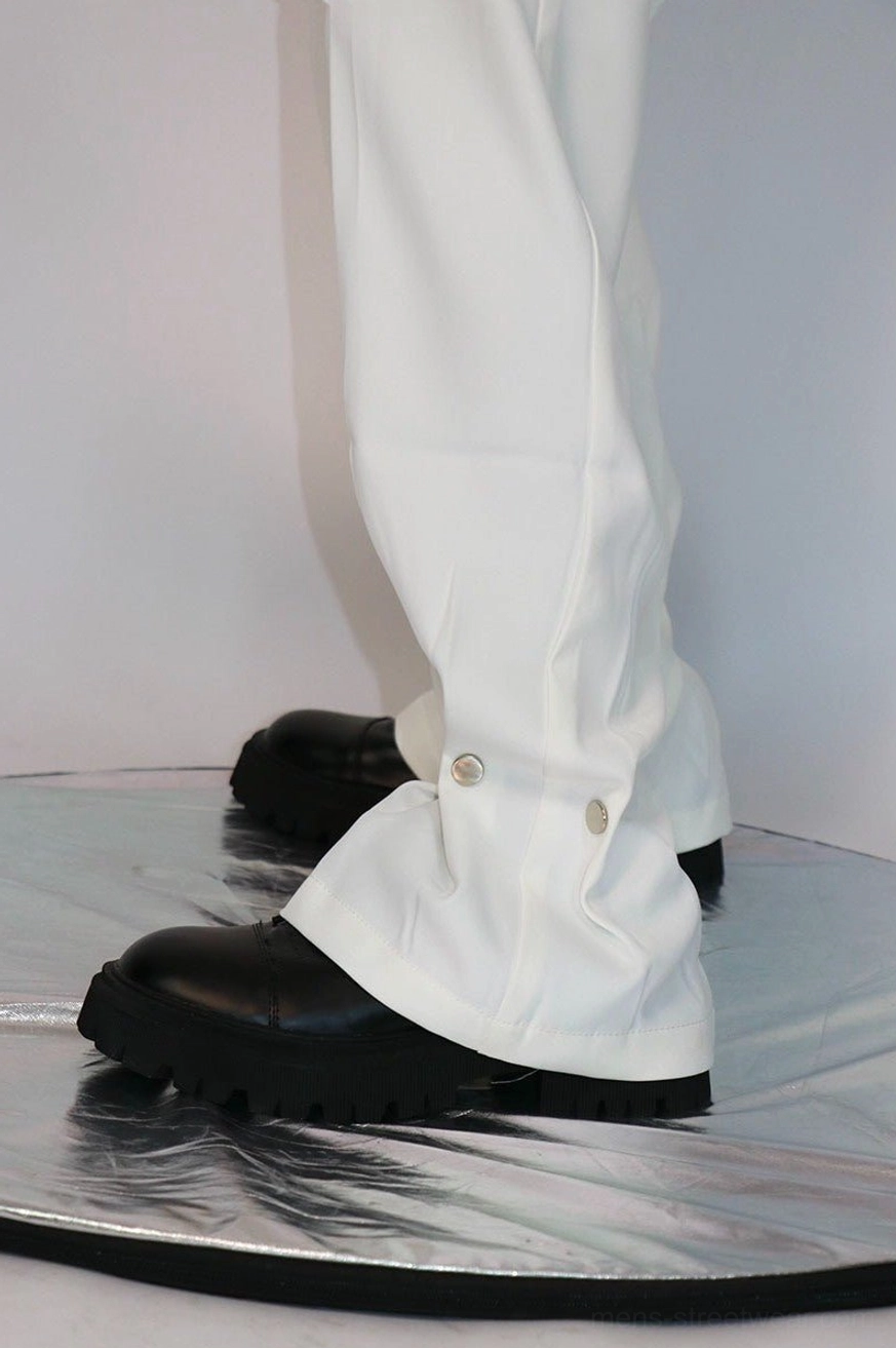 Close-up of white straight-leg streetwear trousers showing tailored front pleats, metal snap fasteners at the cuff, and black platform footwear