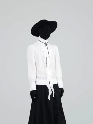 Front full body view of a white Victorian ruffle cravat blouse styled with a black long skirt, wide brim hat, gloves, rings, and dramatic streetwear accessories