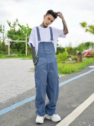 Front full-body view of dusty Haze Blue wide-leg workwear overalls styled with a white T-shirt and white sneakers outdoors