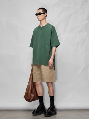 Male model wearing khaki knee-length relaxed streetwear shorts with an oversized dark green pocket tee, black socks, sunglasses, and chunky leather shoes