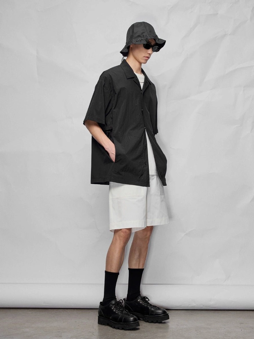Male model in a black short-sleeve work shirt and white knee-length relaxed streetwear shorts with bucket hat and platform shoes