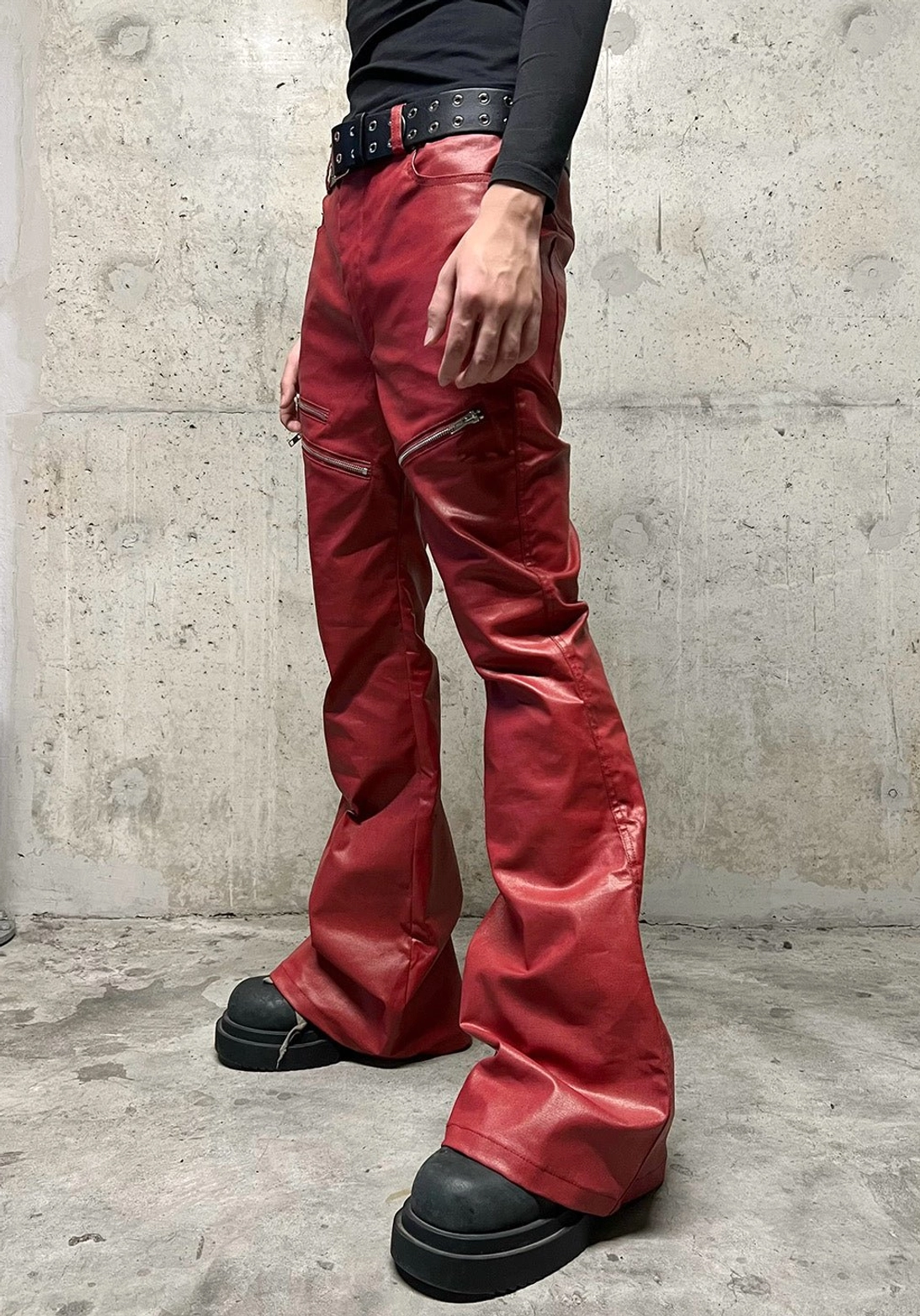 Male model wearing wine red coated flare jeans with fitted high waist, silver zipper hardware, stacked flared legs, black top, studded belt, and platform boots
