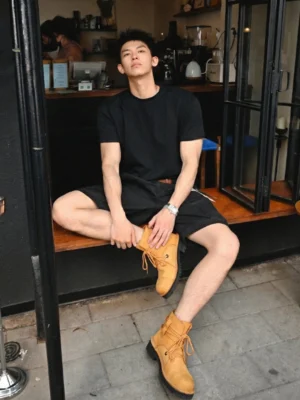 Male model wearing yellow leather utility ankle boots with an all-black casual streetwear outfit at an outdoor cafe