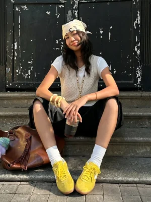 Yellow retro suede-textured streetwear trainers styled with white socks, white tee, knit vest, and black knee-length shorts on an urban street